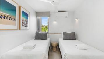 A small, modern room with two single beds, a window with a view of greenery, and a ceiling fan.