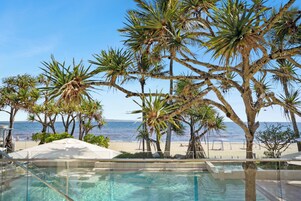 Zona de piscina con agua cristalina, rodeada de palmeras y una playa al fondo.