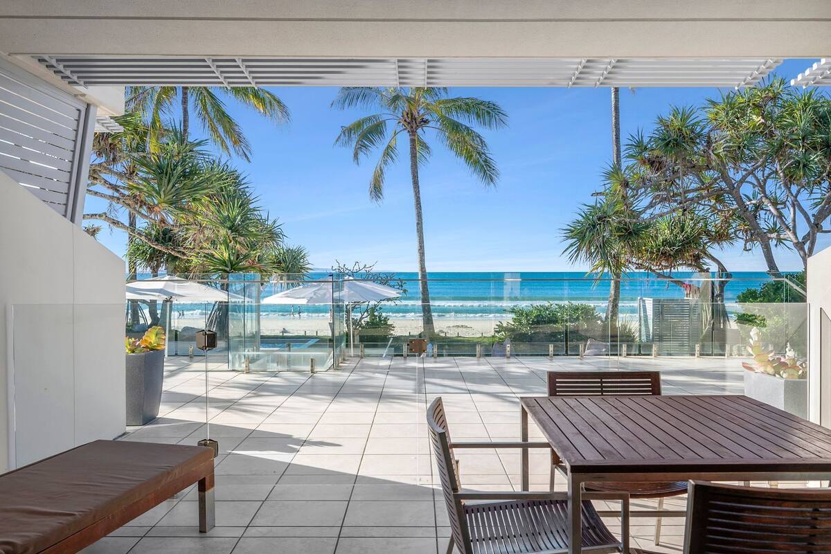 A patio with a view of the beach, a table, chairs, and a bench.