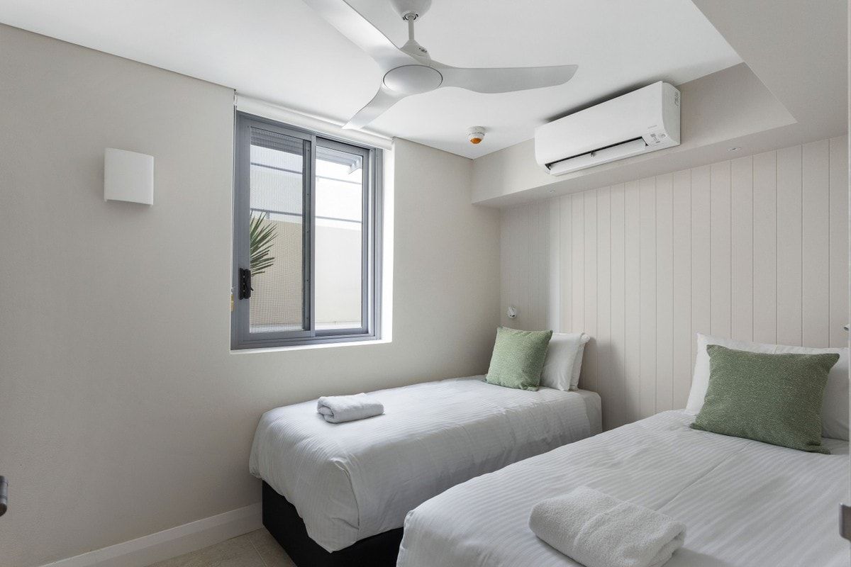 A bedroom with two beds, a ceiling fan, an air conditioning unit, a window with blinds, and a wall-mounted light.