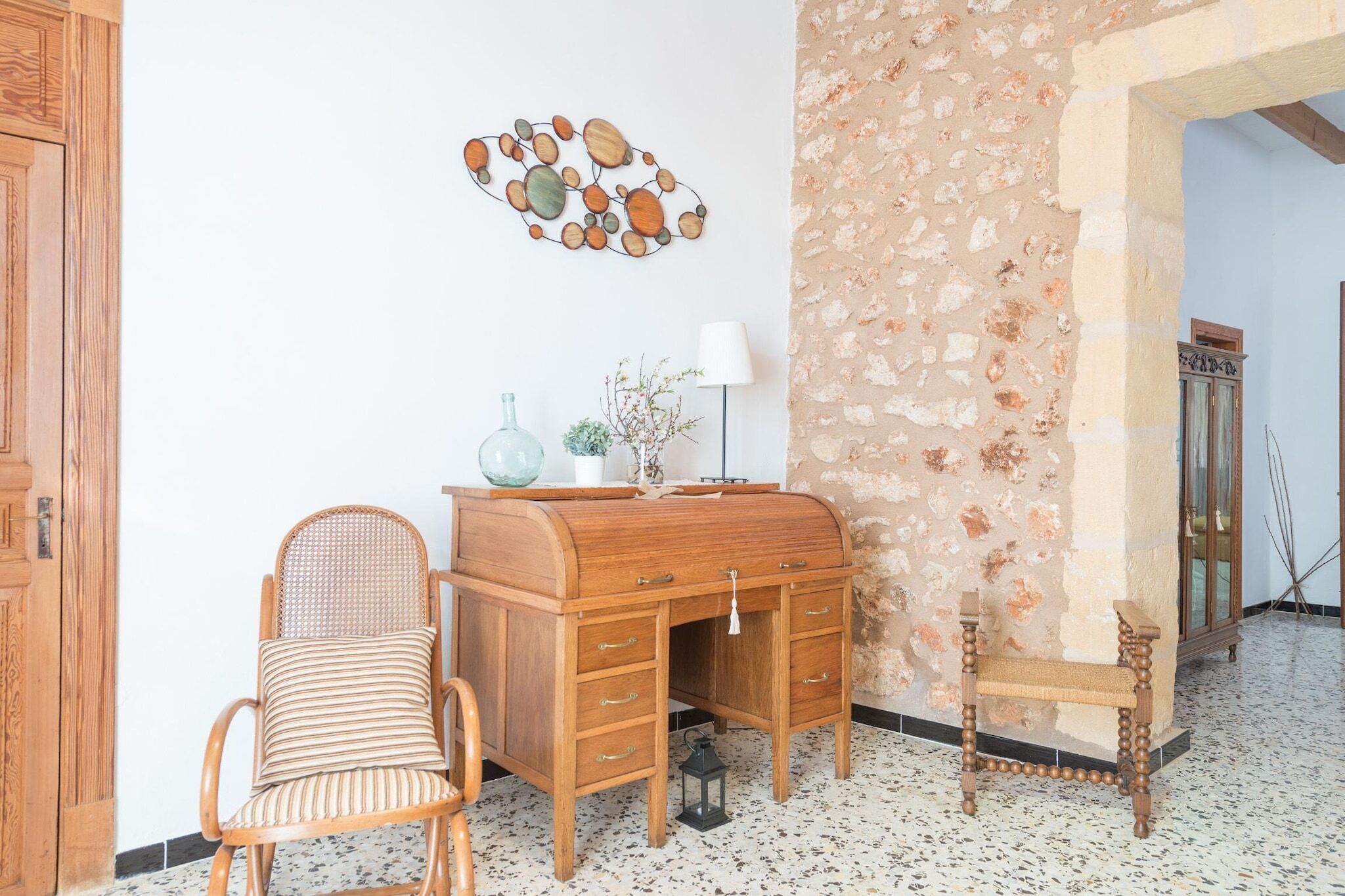 A room with a wooden desk, a wicker chair, a decorative wall piece, and a stone wall.
