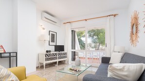 Una sala de estar moderna con una mesa de centro de vidrio, un sofá gris y un sillón amarillo. Se aprecia un mueble blanco con un televisor, un aparador con estampado de cebra y una lámpara blanca. Un ventanal con cortinas blancas ofrece vistas al exterior.