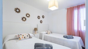 Habitación de hotel con dos camas, una mesita de noche con lámpara, espejos decorativos en la pared y una ventana con cortinas translúcidas.