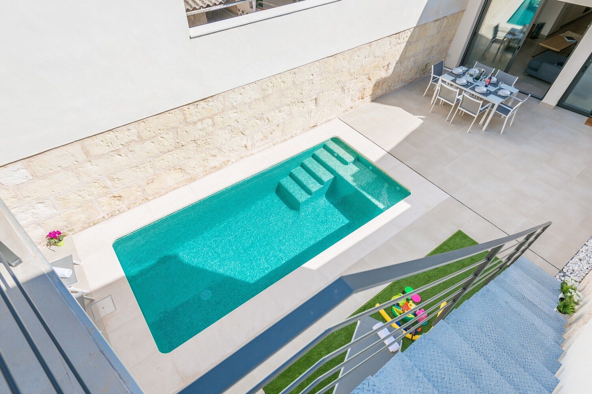 A rooftop pool with a clear blue water feature, a set of stairs leading down to a grassy area with colorful toys, and a dining area with a table and chairs.