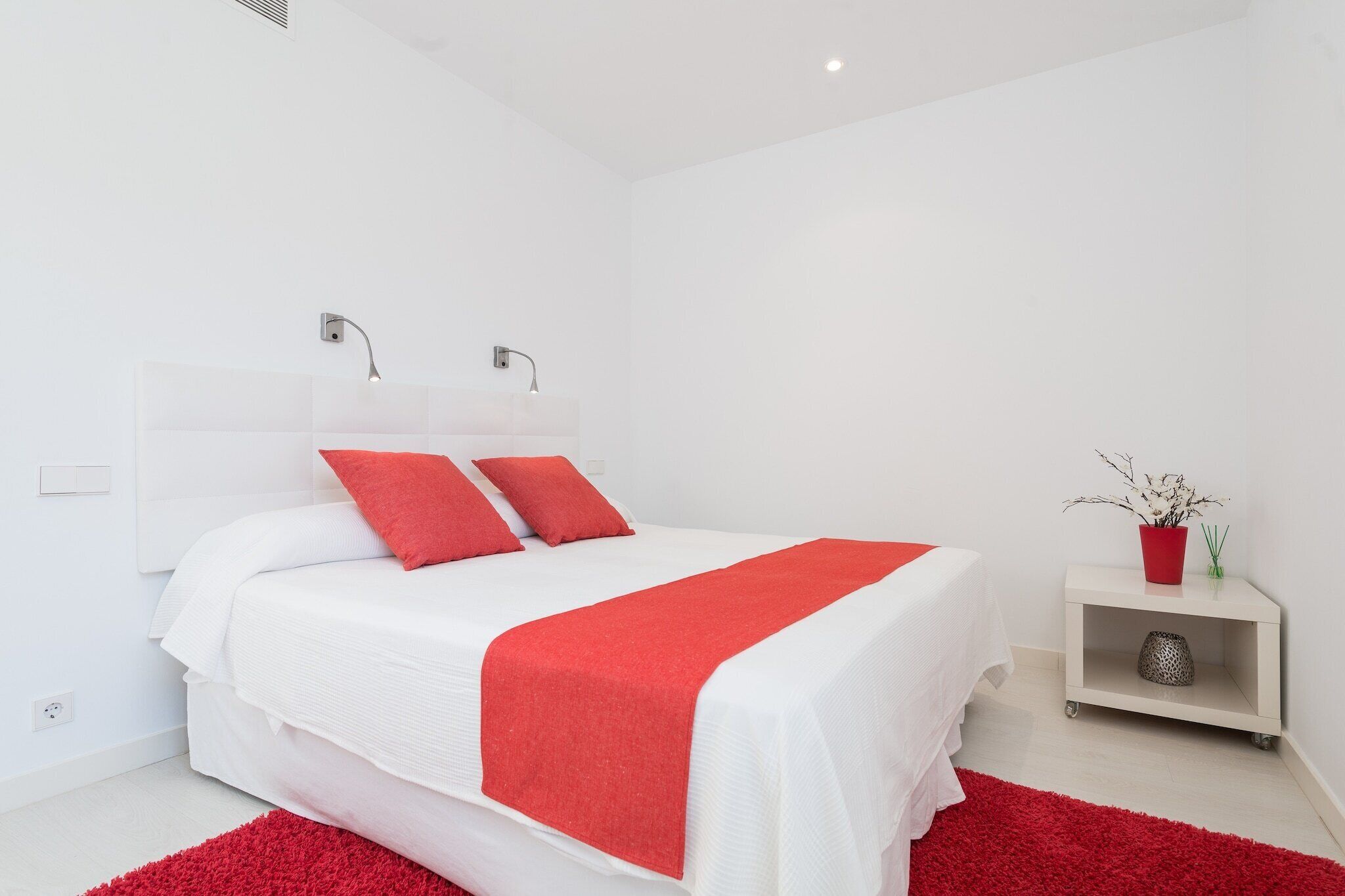Une chambre moderne avec un grand lit, du linge de lit blanc, des coussins rouges et un plaid rouge. On y trouve une petite table de chevet avec un vase et une lampe.