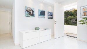 Un intérieur moderne avec une console blanche, trois tableaux encadrés représentant des paysages côtiers, et une porte donnant sur un balcon avec vue sur la verdure et les montagnes.
