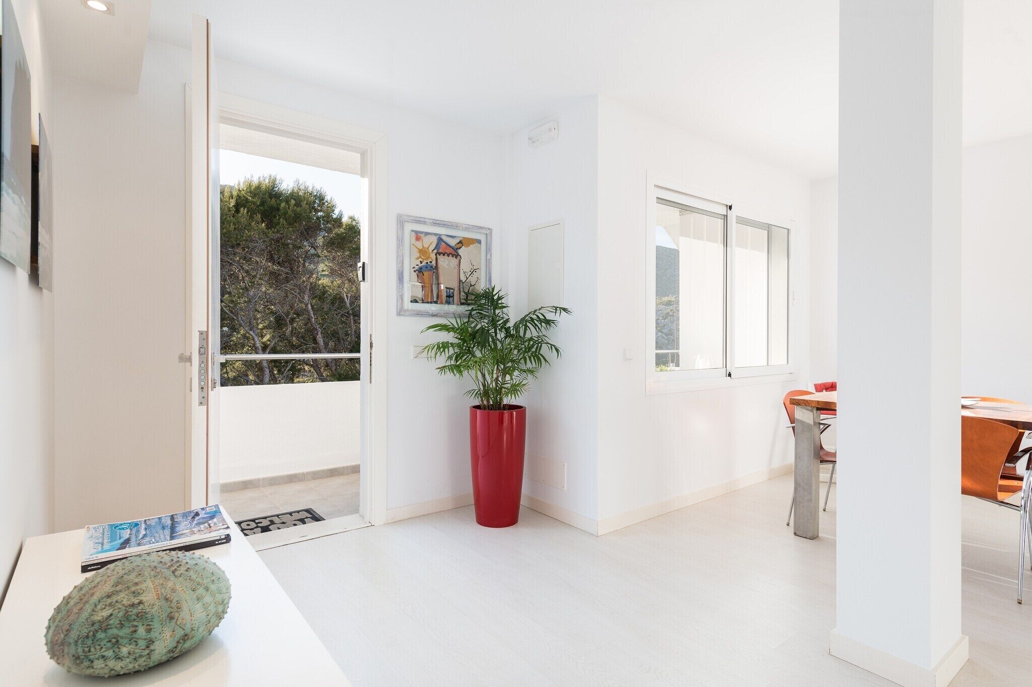 A modern, bright room with a large window, a red potted plant, and a dining area with orange chairs.