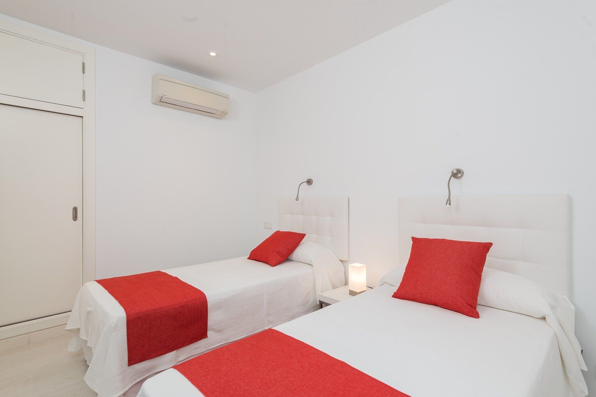 Une chambre d’hôtel avec deux lits, chacun agrémenté d’un oreiller rouge et d’une couverture blanche. On y trouve également une armoire blanche et un appareil de climatisation fixé au mur.