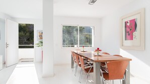 Un espace de repas lumineux et moderne, avec une table en bois et des chaises orange, de grandes fenêtres donnant sur un paysage verdoyant et un décor minimaliste.