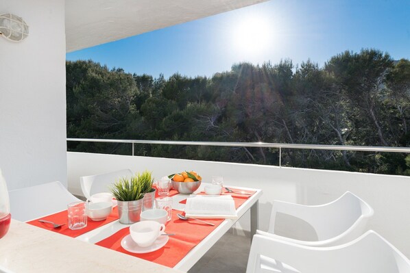 A balcony with a dining table set for two, featuring a bowl of fruit, a potted plant, and a book.