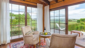 A room with large windows, a wooden ceiling, and a view of a green landscape. There are two chairs and a small table with a bottle and glasses.