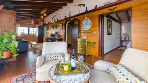 A spacious living area with wooden beams, a round table with a bottle and glasses, and a large clock on the wall.