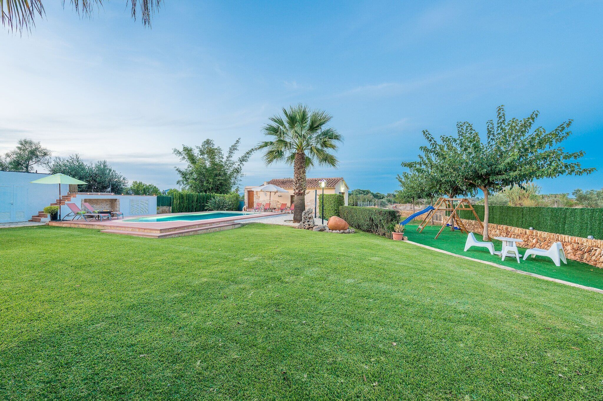 Un patio trasero bien cuidado con piscina, palmera, zona de juegos y una pequeña construcción.