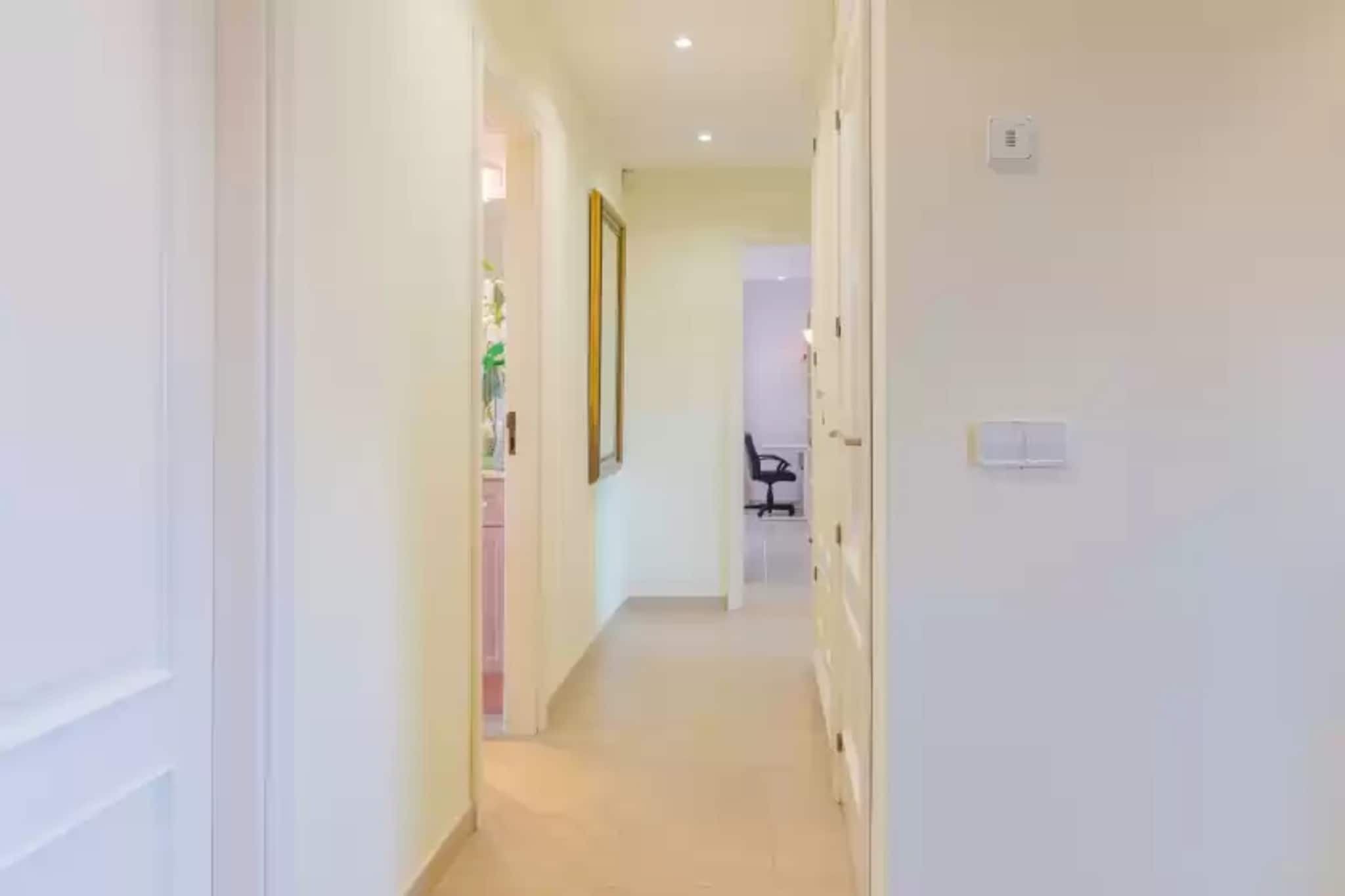 Un couloir étroit aux murs blancs et au sol carrelé, qui mène à une pièce contenant un bureau et une chaise.