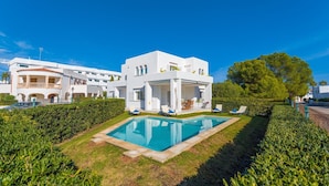 Une villa moderne aux murs blancs, dotée d’une piscine, entourée de haies bien entretenues et surplombant un ciel d’un bleu éclatant.