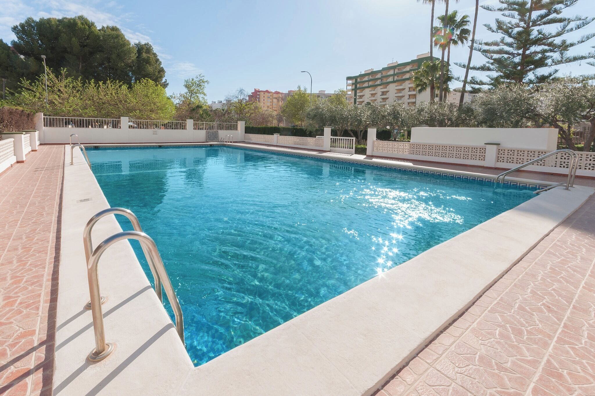 A rectangular swimming pool with clear blue water, surrounded by a tiled deck and a white fence.