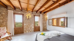 A bedroom with stone walls, wooden beams, a bed with towels, a chair, and a view through a door.
