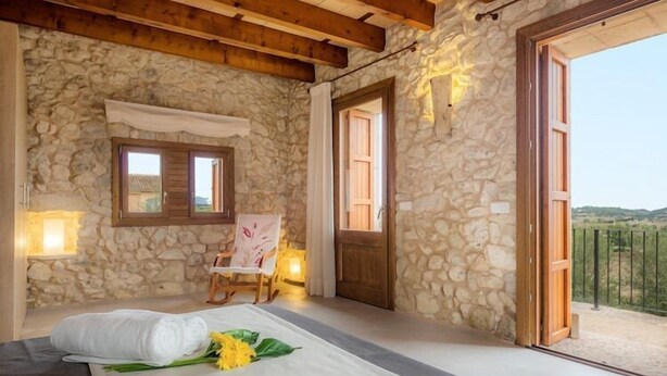 A stone-walled bedroom with a bed, a chair, and a balcony with a view.