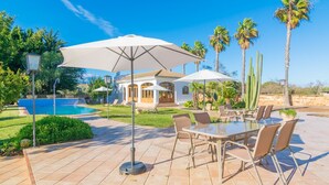 Zona junto a la piscina con mobiliario exterior, palmeras y un edificio al fondo.