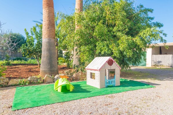 A play area with a small house and a slide, surrounded by trees and a gravel path.