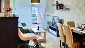 A modern living room with a sofa, coffee table, and dining area. There is a TV and a bookshelf with decorative items.