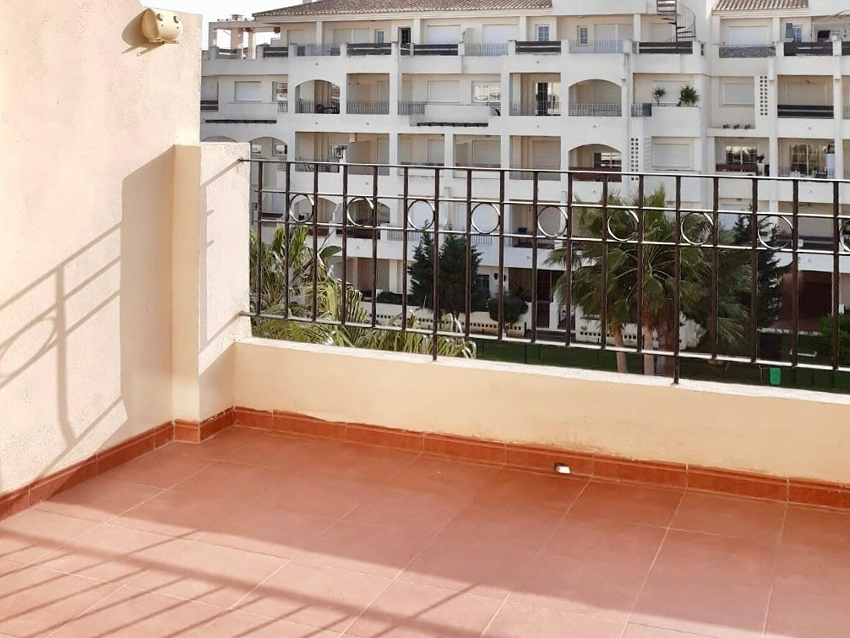 A balcony with a metal railing, a tiled floor, and a view of a multi-story building with balconies and greenery.