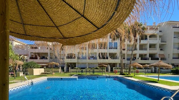 A swimming pool with a clear blue water, surrounded by a covered area with thatched umbrellas, and a multi-story building in the background.
