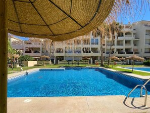 A swimming pool with a clear blue water, surrounded by a covered area with thatched umbrellas, and a multi-story building in the background. - Apartment in Roquetas Near Playa Serena Beach (Roquetas de Mar)