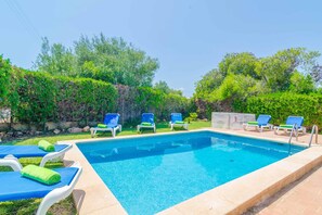 A rectangular swimming pool with clear blue water, surrounded by a tiled deck. There are six lounge chairs with blue cushions and green towels neatly arranged around the pool.