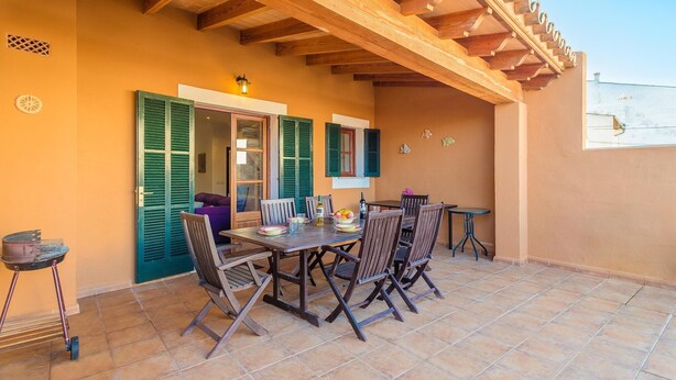 Un patio al aire libre con mesa y sillas de madera, una mesita con lámpara y una ventana con contraventanas verdes.