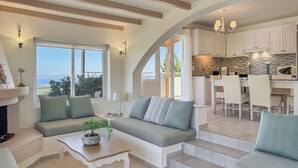 A modern living room with a sofa, a coffee table, and a fireplace. There is a kitchen area with white cabinets and a dining table. - Pano Stalos (Chania)