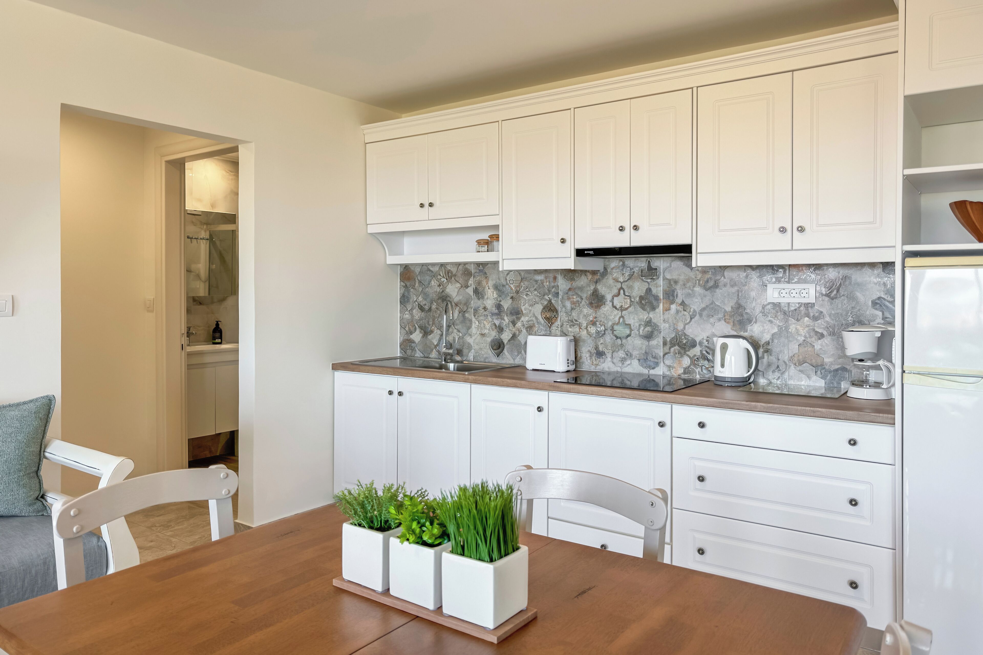 A modern kitchen with white cabinets, a marble backsplash, and a wooden dining table with potted plants.