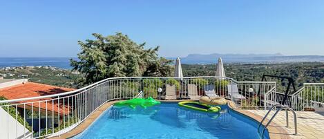 A swimming pool with a view of the sea and mountains, surrounded by a white fence and a red-roofed building.