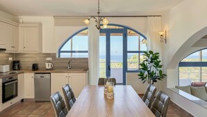 A kitchen with white cabinets, a wooden dining table, and a view of the sea through large windows. - Pano Stalos (Chania)