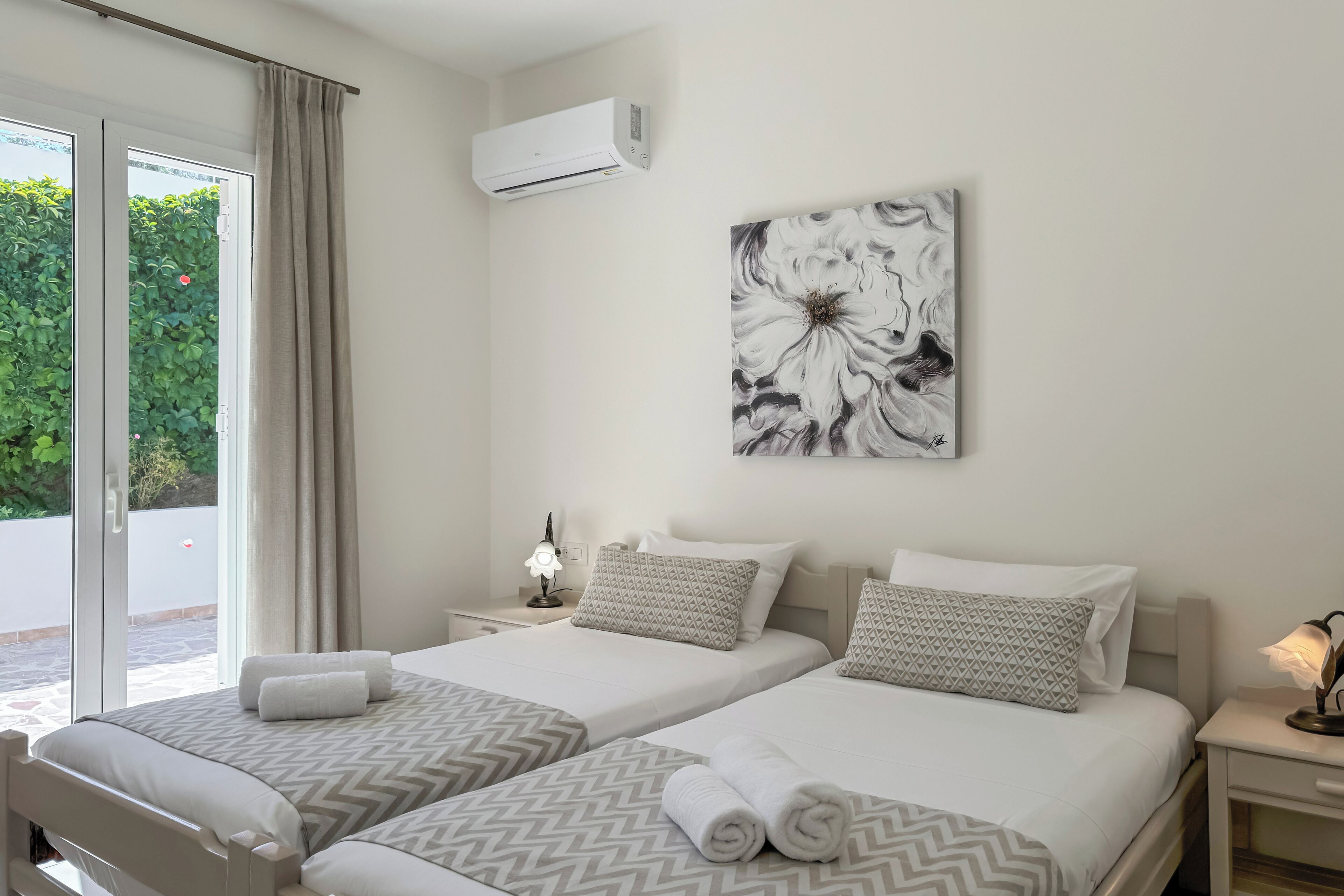 A hotel room with two beds, a large window with curtains, an air conditioning unit, a wall-mounted artwork, and a bedside table with a lamp.