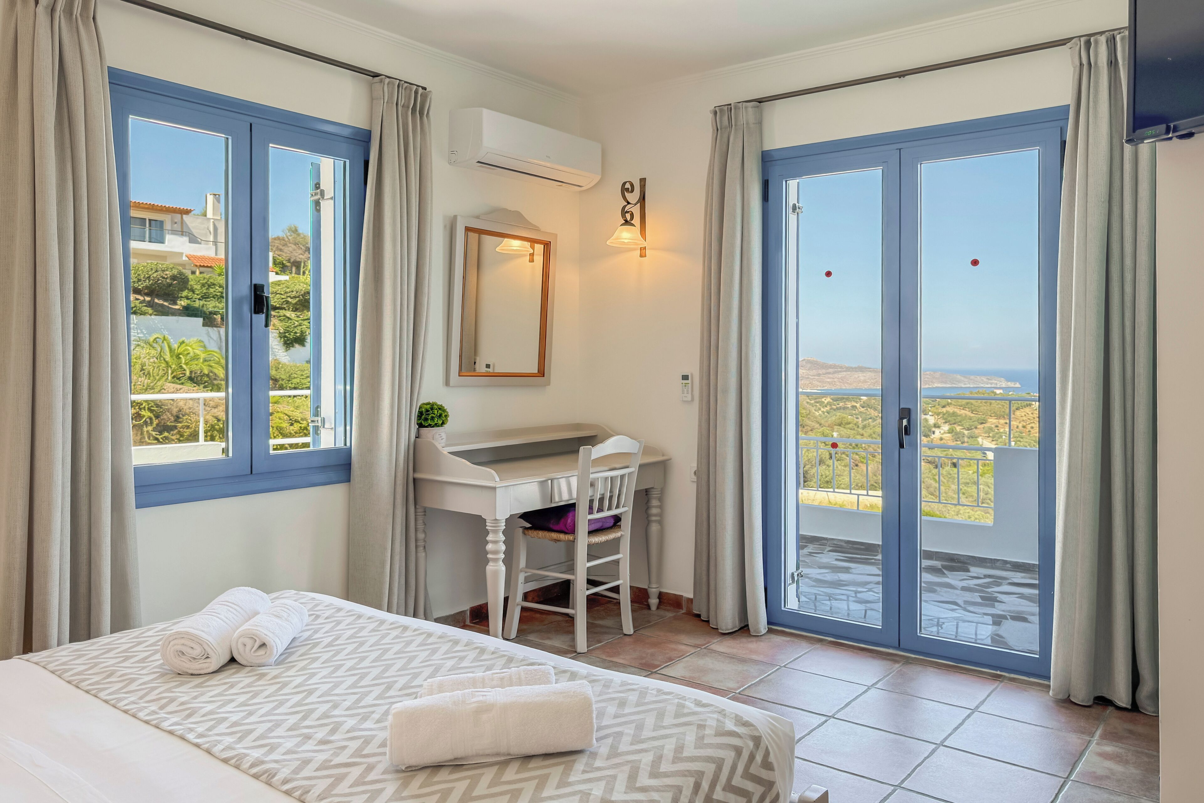 A hotel room with a bed, a desk, and a chair. There is a balcony with a view of the landscape.