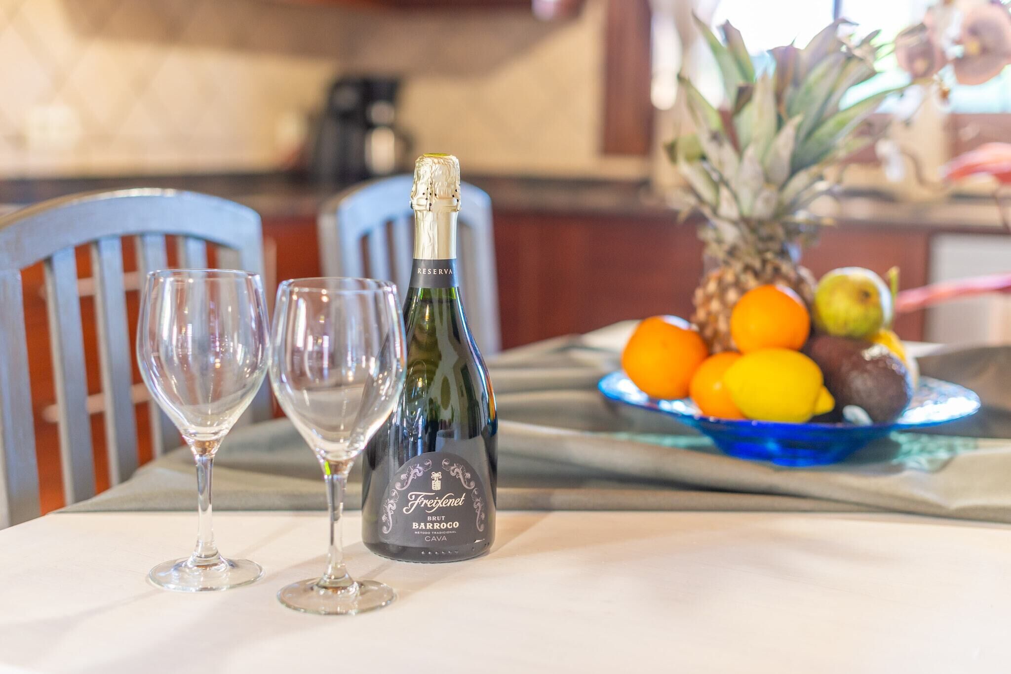 Une table avec deux verres à vin vides, une bouteille de champagne Freixenet et un panier de fruits contenant des oranges et un ananas.