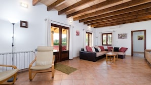 Un salon comprenant un canapé, un fauteuil, une petite table et un radiateur. Il y a des fenêtres et une porte donnant sur l’extérieur.
