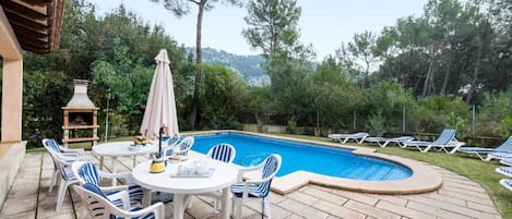 A poolside area with a round table and chairs, a barbecue grill, and lounge chairs.