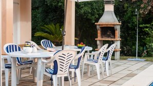 Une terrasse extérieure avec des chaises blanches et une table, un four en briques et un grand parasol.