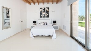 Une chambre avec un grand lit, des murs blancs, des poutres en bois au plafond, une grande fenêtre et un tableau au-dessus du lit.