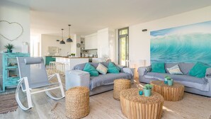 Un salón moderno con un sofá gris, un sillón mecedora y una mesa de centro de madera. Se ve una cocina al fondo con armarios blancos y un ventanal grande.