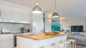 Una cocina moderna con armarios blancos, encimera de madera y una isla central con taburetes. Se observa un cuadro grande de una ola en la pared, una televisión y una lámpara a la derecha.