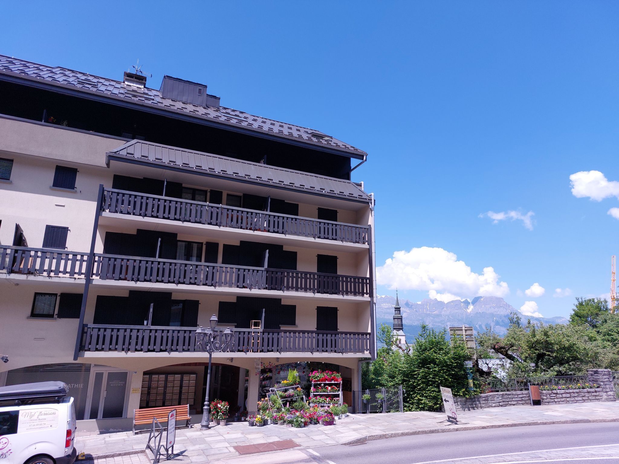 A multi-story building with balconies, a flower shop on the ground floor, and a mountain view in the background.
