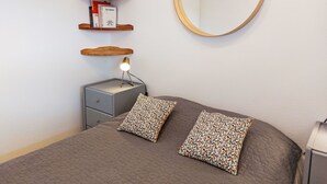 Un dormitorio con cama, dos almohadones estampados, lámpara de noche, mesita pequeña, espejo y estantes de madera con libros y una botella.