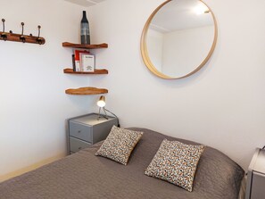 Ein Schlafzimmer mit einem Bett, zwei gemusterten Kissen, einer Nachttischlampe, einem kleinen Nachttisch, einem Spiegel und Holzregalen mit Büchern und einer Flasche.