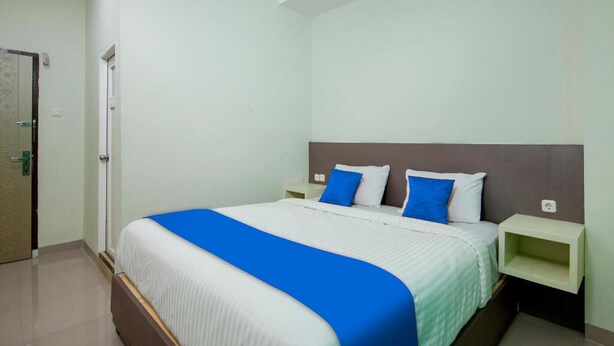 A hotel room with a large bed, white and blue bedding, a wooden headboard, and a nightstand with a lamp. - Hotel Wisata Samosir By Helocus (Samosir)