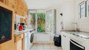 Cozinha compacta com armários de madeira, máquina de lavar, cafeteira e vista para o verde lá fora.
