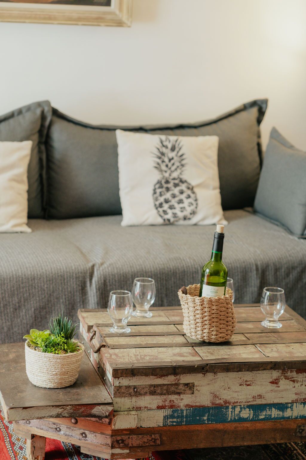 Een gezellige woonkamer met een grijze bank, kussens met ananasprint, een rustieke salontafel met een fles wijn en glazen, en een kleine potplant.