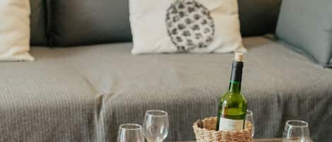 A cozy living room with a grey sofa, pineapple-themed pillows, a rustic coffee table with a bottle of wine and glasses, and a small potted plant.